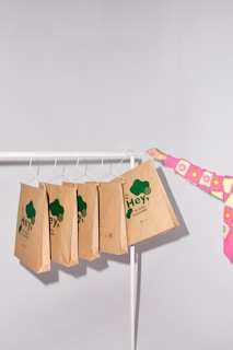 A minimalist setting featuring several brown paper bags with green text hanging on a white clothing rack. The bags have eco-friendly slogans emphasizing recyclability. A person wearing a colorful pink and yellow sweater is holding one of the bags.