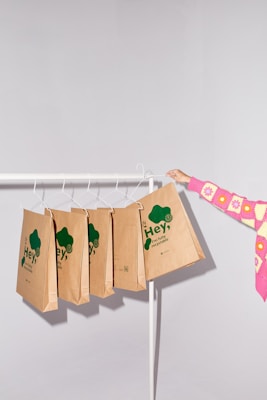 A minimalist setting featuring several brown paper bags with green text hanging on a white clothing rack. The bags have eco-friendly slogans emphasizing recyclability. A person wearing a colorful pink and yellow sweater is holding one of the bags.