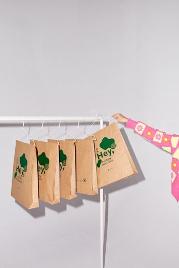 A minimalist setting featuring several brown paper bags with green text hanging on a white clothing rack. The bags have eco-friendly slogans emphasizing recyclability. A person wearing a colorful pink and yellow sweater is holding one of the bags.