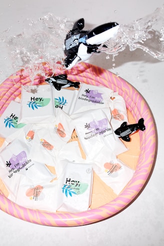Several biodegradable product packages are arranged in a colorful inflatable pool. An inflatable whale toy is splashing water, creating a dynamic and playful scene. The packages are designed with various pastel colors, indicating different features or scents.