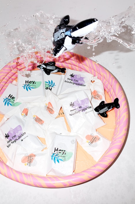 Several biodegradable product packages are arranged in a colorful inflatable pool. An inflatable whale toy is splashing water, creating a dynamic and playful scene. The packages are designed with various pastel colors, indicating different features or scents.