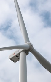 A close-up view of a wind turbine against a background of partly cloudy skies. The image focuses on the rotor and blades of the turbine, which appear to be made of smooth, white material. A logo with lettering is visible on the turbine's nacelle.