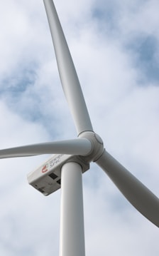A close-up view of a wind turbine against a background of partly cloudy skies. The image focuses on the rotor and blades of the turbine, which appear to be made of smooth, white material. A logo with lettering is visible on the turbine's nacelle.