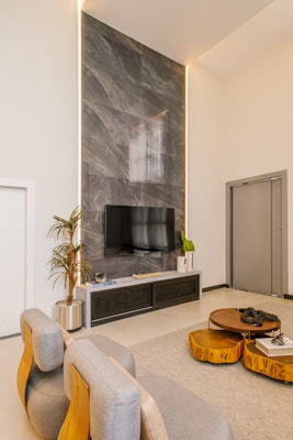 A modern living room features a large, flat-screen television mounted against a tall, gray marble accent wall. Below the TV is a sleek console table with decorative items and a potted plant. The room includes stylish chairs with wooden accents, and a set of round wooden coffee tables. Neutral tones dominate the decor, complemented by a light-colored area rug and minimalistic design elements.