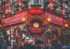 A bustling street scene captures a vibrant pub with bright red exterior and ornate detailing. People are gathered outside, some taking photos, while hanging plants and festive lights adorn the building. The pub's signage reads 'Waxy's Little Sister', adding character to the lively atmosphere.