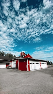 A red building with multiple white garage doors under a partly cloudy sky. The structure sits on a paved surface and has a backdrop of trees on the left side.