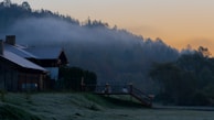 A serene landscape featuring houses with sloped roofs situated near a gently sloping hillside. Mist envelops the distant forested hills, creating a soft transition into the early morning sky, which is painted with hues of orange and blue.