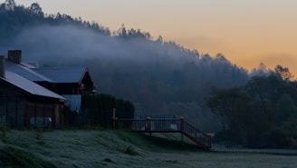 A serene landscape featuring houses with sloped roofs situated near a gently sloping hillside. Mist envelops the distant forested hills, creating a soft transition into the early morning sky, which is painted with hues of orange and blue.