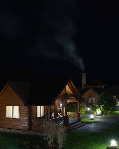 A wooden cabin surrounded by a dimly lit garden, with light coming from windows and garden lamps. Smoke is rising from a chimney in the background, suggesting a cool night. The atmosphere is calm and serene, with neatly trimmed grass and a stone pathway.
