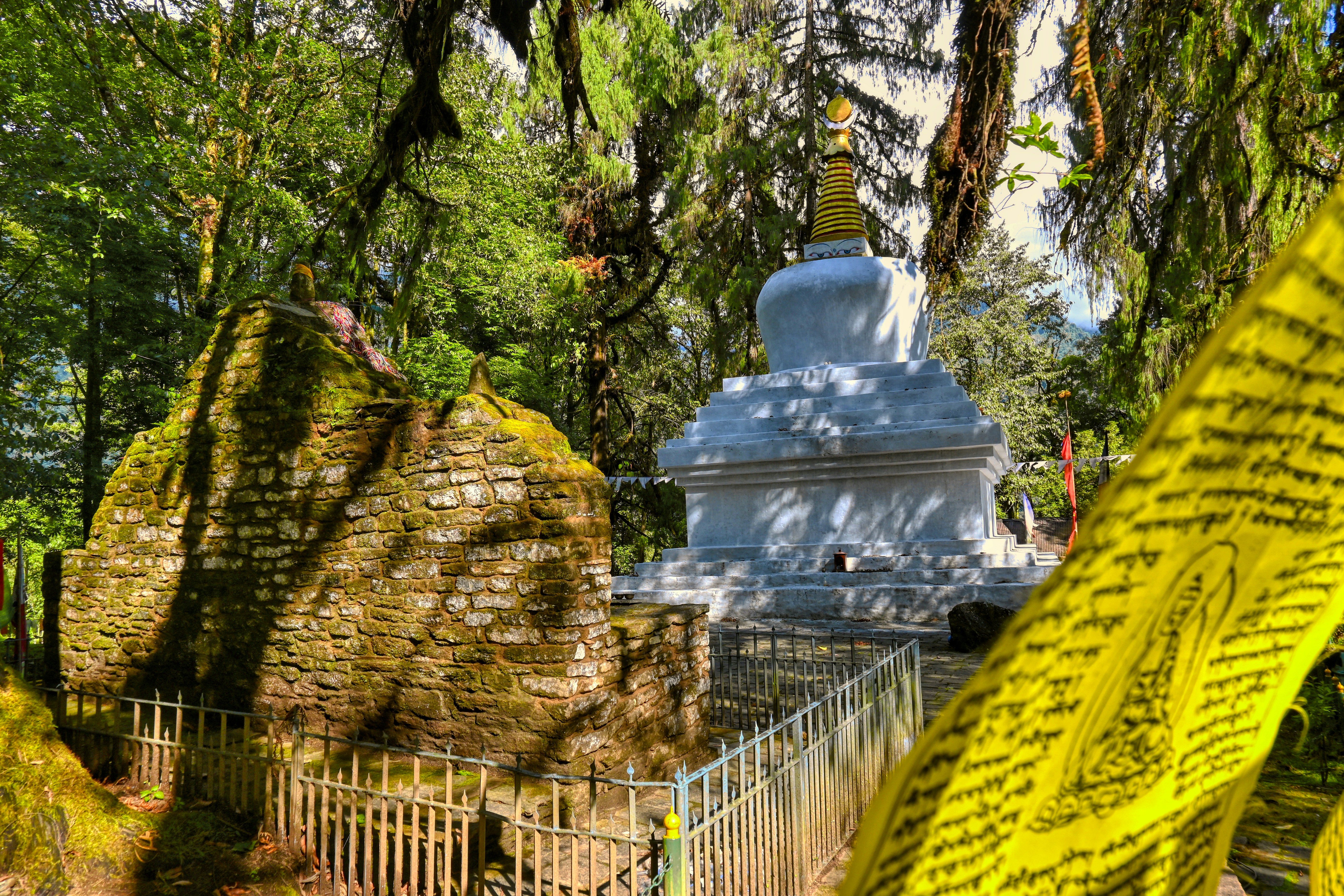 A whitewashed chorten beside the moss-covered stone coronation throne at Norbugang in Yuksom, yellow prayer flags in front