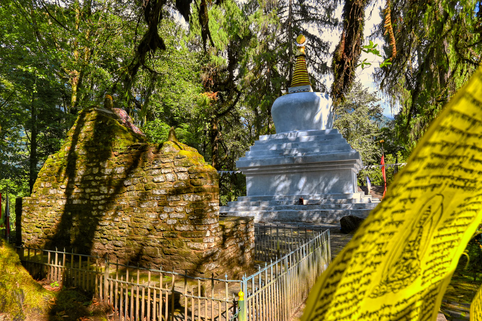 A whitewashed chorten beside the moss covered stone coronation throne at Norbugang in Yuksom, yellow prayer flags in front