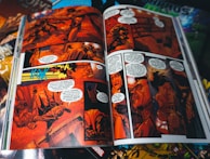 A comic book is open to a colorful, action-packed spread. The pages are filled with detailed illustrations and dialogue bubbles, prominently featuring characters in dramatic scenes and dynamic poses. The artwork is vibrant, with a focus on red and orange hues that create an intense atmosphere.