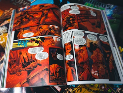 A comic book is open to a colorful, action-packed spread. The pages are filled with detailed illustrations and dialogue bubbles, prominently featuring characters in dramatic scenes and dynamic poses. The artwork is vibrant, with a focus on red and orange hues that create an intense atmosphere.