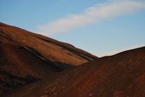 A landscape featuring rolling hills with a reddish-brown hue under a clear blue sky. The sunlight casts soft shadows, creating a serene and dramatic effect.