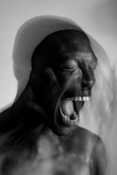 A black and white photograph captures a person with an intense expression, possibly screaming or shouting. The motion blur effect adds a dynamic and emotional element to the image, emphasizing movement and intensity. The background is simple, focusing attention on the person.