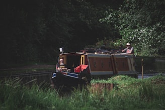 A narrow canal boat navigates through a lush, green waterway. Two people are on board, with one steering at the rear and another sitting at the front. The boat is adorned with plants and appears to be moving slowly along the canal. Dense trees and foliage surround the water, casting shadows over the scene.