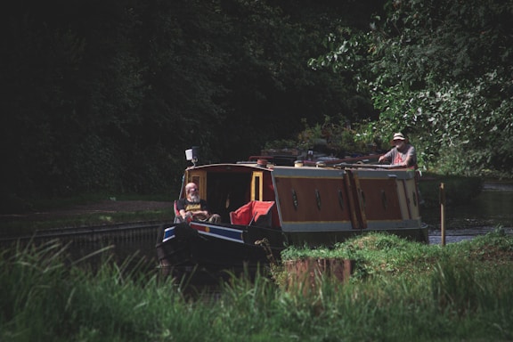 A narrow canal boat navigates through a lush, green waterway. Two people are on board, with one steering at the rear and another sitting at the front. The boat is adorned with plants and appears to be moving slowly along the canal. Dense trees and foliage surround the water, casting shadows over the scene.