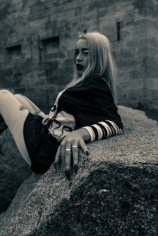 A person with long blonde hair and dark lipstick is leaning back on a large rock. They are wearing a dark outfit with a design on the chest and have rings on their fingers. The background features a stone wall with an industrial, urban feel.