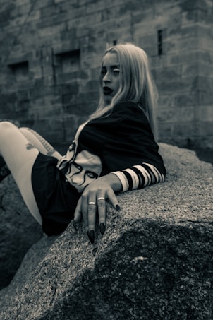 A person with long blonde hair and dark lipstick is leaning back on a large rock. They are wearing a dark outfit with a design on the chest and have rings on their fingers. The background features a stone wall with an industrial, urban feel.