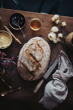 A rustic wooden table showcasing jars of colorful homemade jams and jellies next to a fresh loaf of bread dusted with flour.