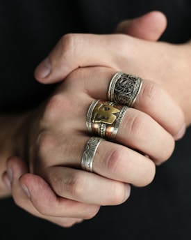 Hands are clasped together, displaying several ornate rings with intricate designs. The rings vary in size and style, featuring detailed patterns in silver and gold tones.