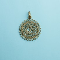 A bronze-colored pendant with intricate mandala patterns and an Om symbol in the center rests on a light blue background.