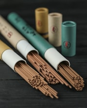 A collection of incense sticks partially pulled out of cylindrical cardboard containers, with more containers standing upright in the background on a dark wooden surface.