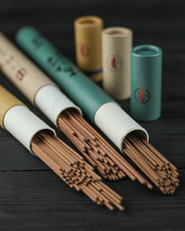 A collection of incense sticks partially pulled out of cylindrical cardboard containers, with more containers standing upright in the background on a dark wooden surface.
