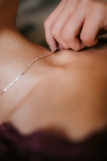 A close-up view of a hand holding a delicate silver necklace against a person's skin. The focus is on the intricate details of the jewelry and the gentle touch of the fingers.