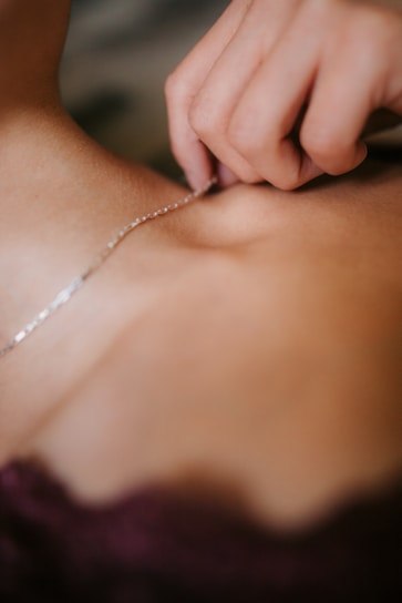 A close-up view of a hand holding a delicate silver necklace against a person's skin. The focus is on the intricate details of the jewelry and the gentle touch of the fingers.