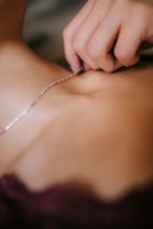 A close-up view of a hand holding a delicate silver necklace against a person's skin. The focus is on the intricate details of the jewelry and the gentle touch of the fingers.