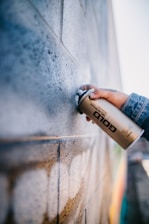 A person is holding a gold spray paint can near a textured brick wall. The wall appears to have been partially sprayed with a light color, creating an artistic and urban feel. The person is wearing a denim jacket, and their hand is prominently focused.