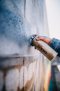 A person is holding a gold spray paint can near a textured brick wall. The wall appears to have been partially sprayed with a light color, creating an artistic and urban feel. The person is wearing a denim jacket, and their hand is prominently focused.