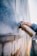 A person is holding a gold spray paint can near a textured brick wall. The wall appears to have been partially sprayed with a light color, creating an artistic and urban feel. The person is wearing a denim jacket, and their hand is prominently focused.