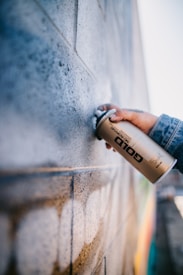 A person is holding a gold spray paint can near a textured brick wall. The wall appears to have been partially sprayed with a light color, creating an artistic and urban feel. The person is wearing a denim jacket, and their hand is prominently focused.