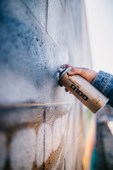 A person is holding a gold spray paint can near a textured brick wall. The wall appears to have been partially sprayed with a light color, creating an artistic and urban feel. The person is wearing a denim jacket, and their hand is prominently focused.