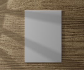 A blank white piece of paper is placed on a wooden surface with a textured grain pattern. Soft shadows fall across the paper, adding depth to the flat layout.