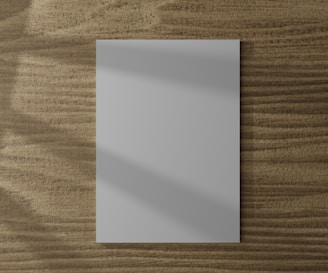 A blank white piece of paper is placed on a wooden surface with a textured grain pattern. Soft shadows fall across the paper, adding depth to the flat layout.