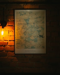A map of Białystok hangs on a brick wall, illuminated warmly by a nearby bare light bulb. The map, set in a simple black frame, shows various roads and geographical features, with an artistic color palette that contrasts against the rustic texture of the bricks.