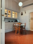 A cozy kitchen corner with a round table surrounded by four wooden chairs. The floor is made of reddish hexagonal tiles, and there is a white cabinet with glass doors above the counter. The cabinet displays various jars and kitchen items. A spherical light hangs from the ceiling, and some decorative items are on the walls.
