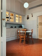 A cozy kitchen corner with a round table surrounded by four wooden chairs. The floor is made of reddish hexagonal tiles, and there is a white cabinet with glass doors above the counter. The cabinet displays various jars and kitchen items. A spherical light hangs from the ceiling, and some decorative items are on the walls.