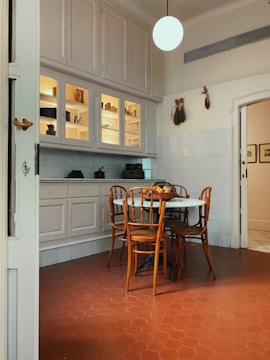 A cozy kitchen corner with a round table surrounded by four wooden chairs. The floor is made of reddish hexagonal tiles, and there is a white cabinet with glass doors above the counter. The cabinet displays various jars and kitchen items. A spherical light hangs from the ceiling, and some decorative items are on the walls.