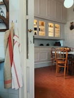 A cozy kitchen interior featuring a rustic design with wooden chairs around a round table on a red hexagonal tile floor. The walls have white cabinets with some glass doors, displaying crockery and kitchen items. On the left, a set of linen towels is hanging, and there are decorations like a blue and white ceramic plate on the wall.