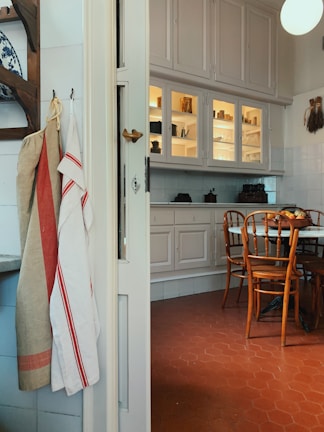 A cozy kitchen interior featuring a rustic design with wooden chairs around a round table on a red hexagonal tile floor. The walls have white cabinets with some glass doors, displaying crockery and kitchen items. On the left, a set of linen towels is hanging, and there are decorations like a blue and white ceramic plate on the wall.