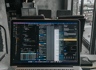 A laptop displaying code on the screen with various open applications is placed on a table in a modern workspace. The background features a concrete wall and windows offering a view of a neighboring building and street below.