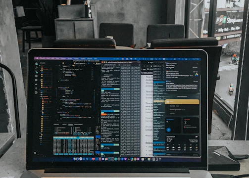A laptop displaying code on the screen with various open applications is placed on a table in a modern workspace. The background features a concrete wall and windows offering a view of a neighboring building and street below.