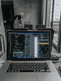 A laptop displaying code on the screen with various open applications is placed on a table in a modern workspace. The background features a concrete wall and windows offering a view of a neighboring building and street below.