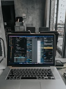 A laptop displaying code on the screen with various open applications is placed on a table in a modern workspace. The background features a concrete wall and windows offering a view of a neighboring building and street below.