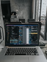 A laptop displaying code on the screen with various open applications is placed on a table in a modern workspace. The background features a concrete wall and windows offering a view of a neighboring building and street below.