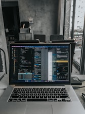 A laptop displaying code on the screen with various open applications is placed on a table in a modern workspace. The background features a concrete wall and windows offering a view of a neighboring building and street below.
